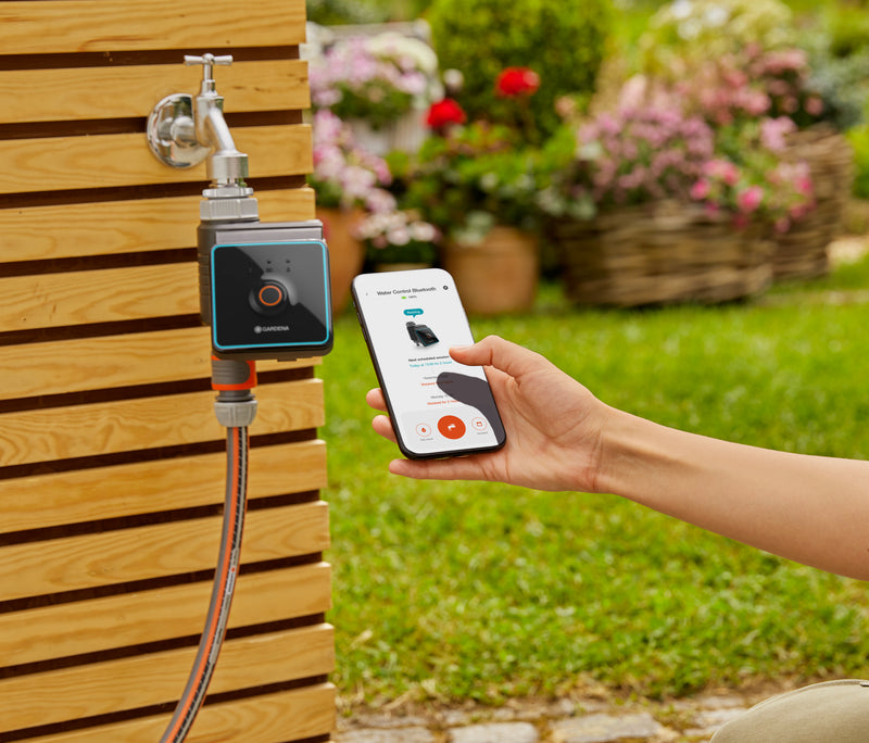 Gardena Bluetooth water timer on an outdoor faucet beside a phone showing the timer app